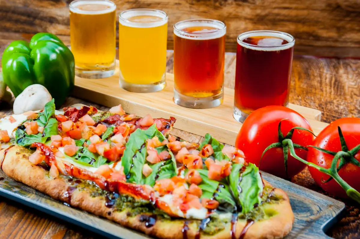 A flatbread pizza topped with vegetables and a flight of beers in the background.