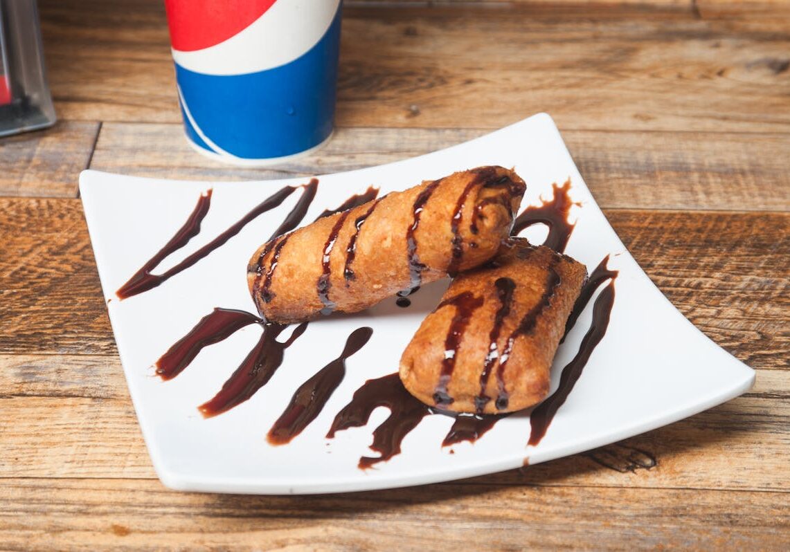 asset 57 Two chocolate-drizzled pastries on a white plate with a soda cup in the background.
